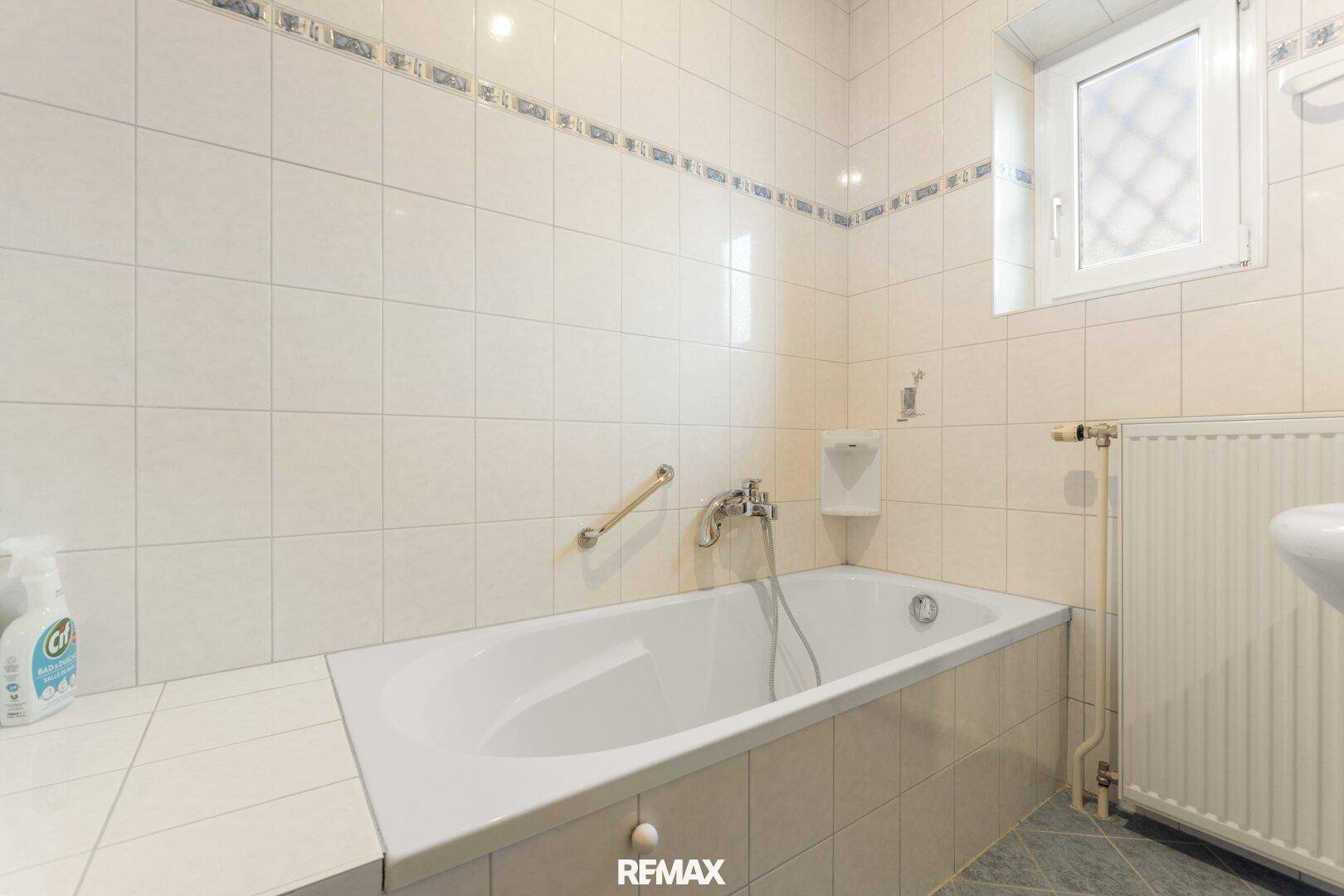 Gefliestes Badezimmer mit Badewanne, Fenster und Handlauf.