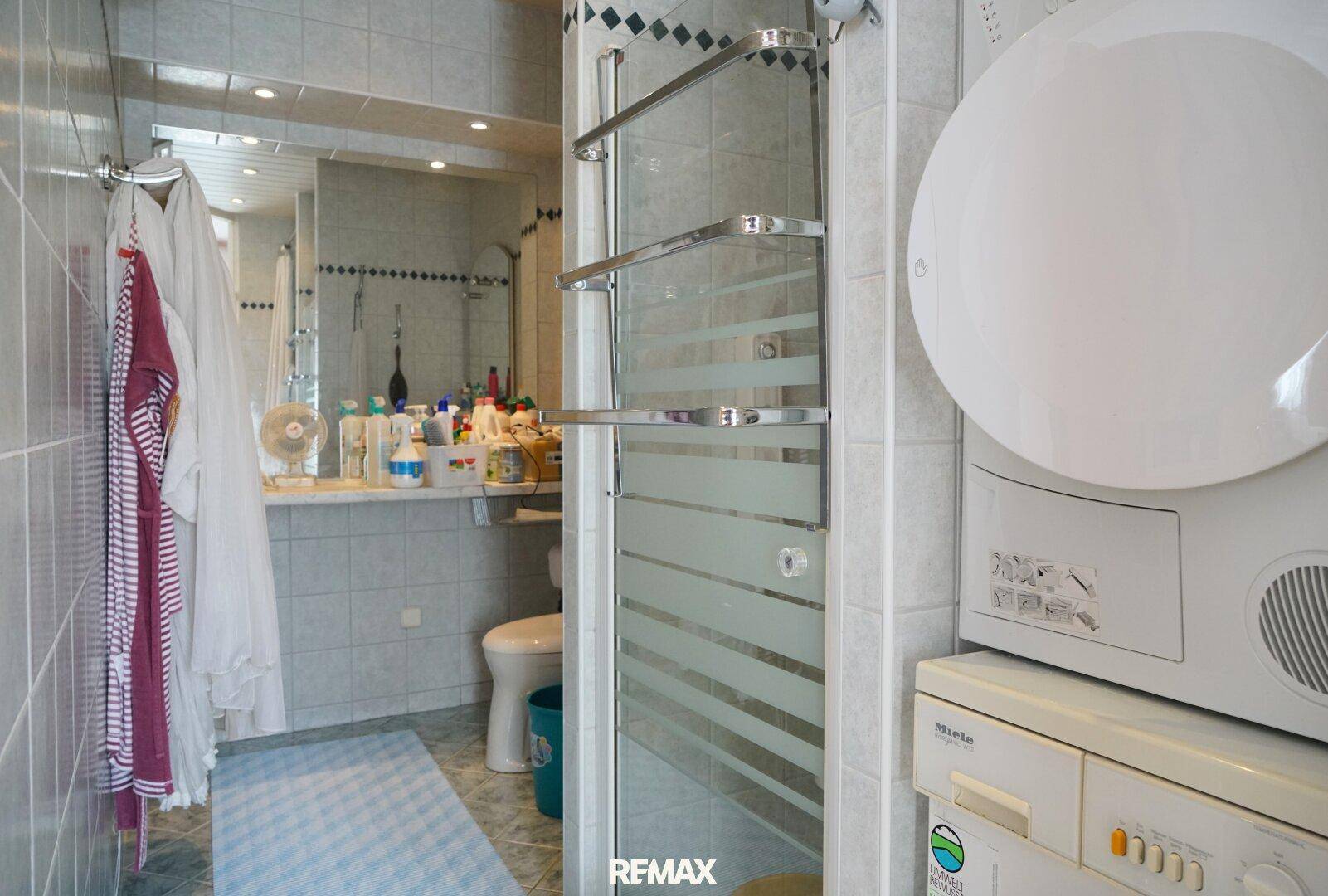 Funktionales Badezimmer mit Dusche, Waschmaschine, Trockner und hellen Fliesen.