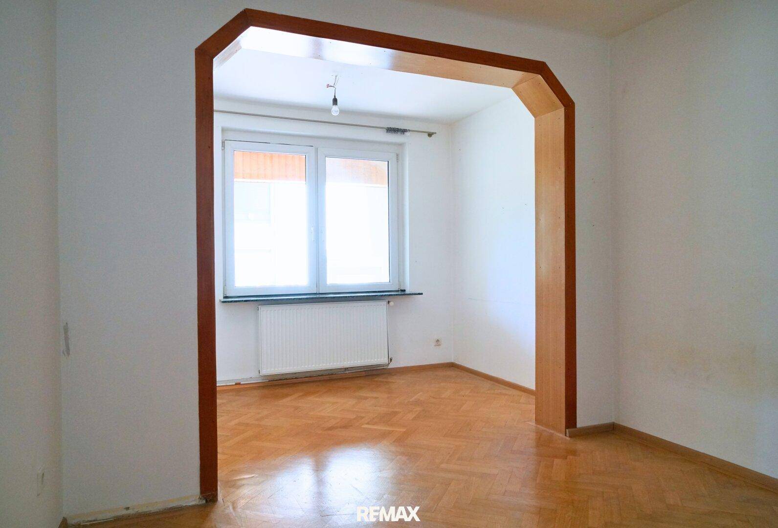 Leerer Raum mit Fischgrätparkett, großem Fenster und Heizkörper, bereit zur individuellen Gestaltung.