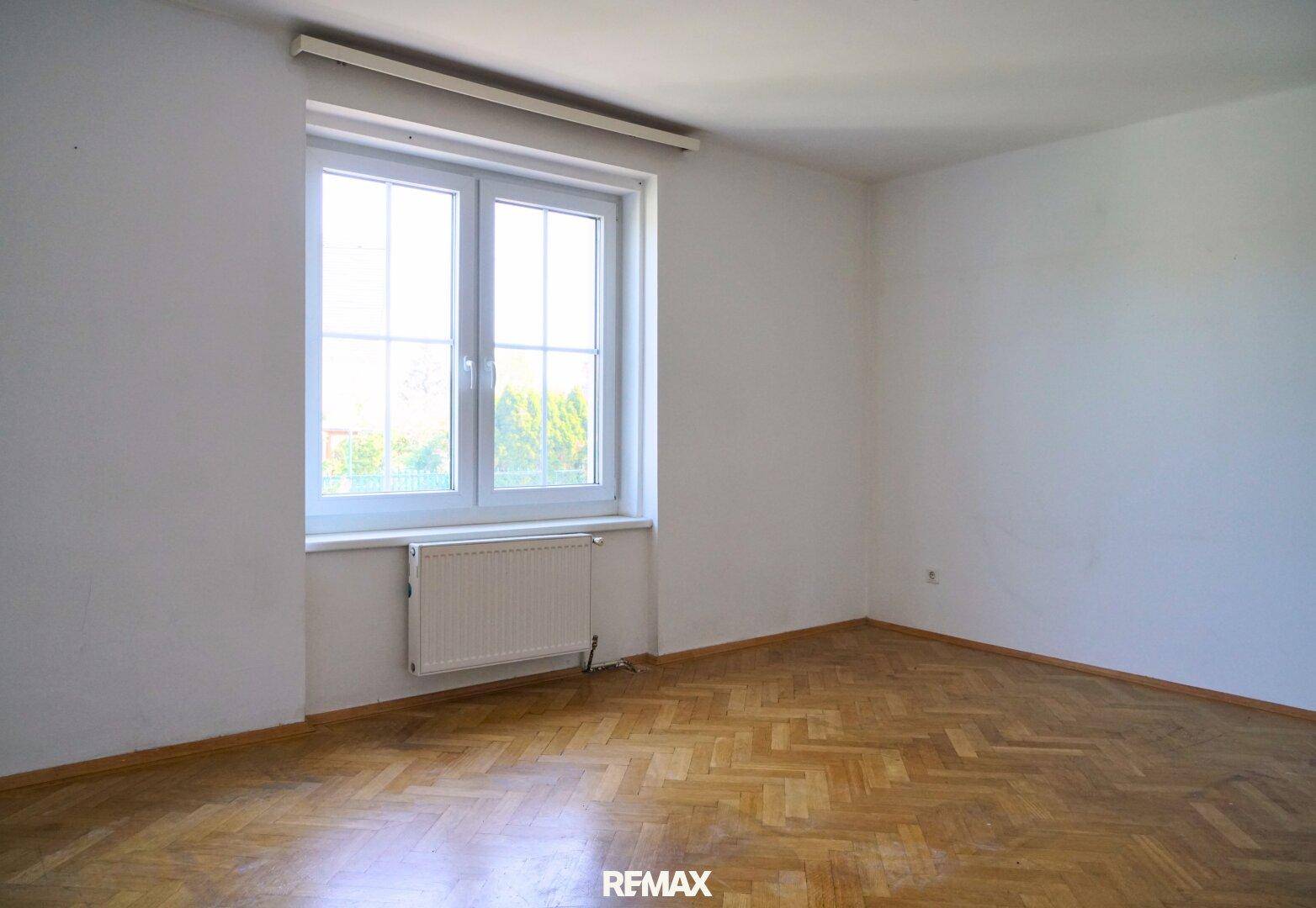 Leeres Zimmer mit Parkettboden, weißen Wänden und großem Fenster.
