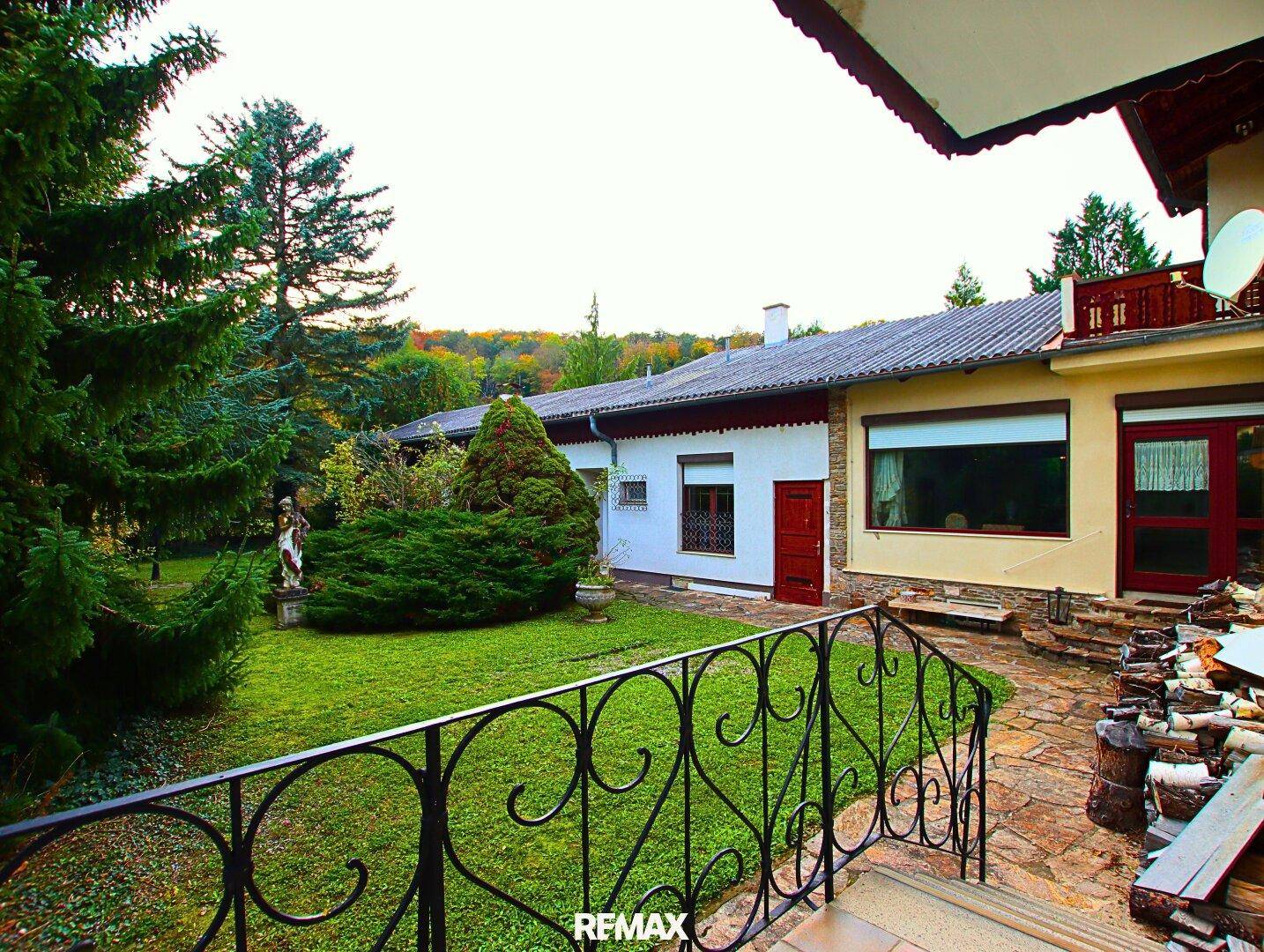 Blick vom Garten auf das Haus mit einer gepflasterten Terrasse, schmiedeeisernem Geländer und altem Baumbestand.
