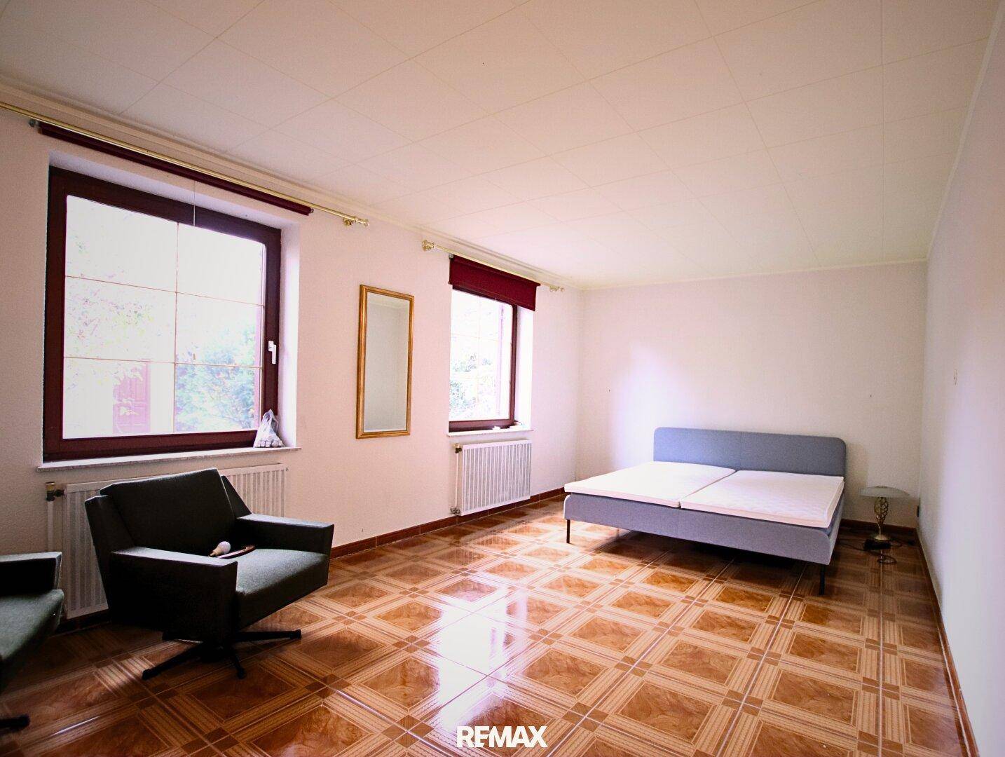Schlafzimmer mit gemustertem Fliesenboden, einem Bett, Sessel und zwei Fenstern, die viel Licht hereinlassen.