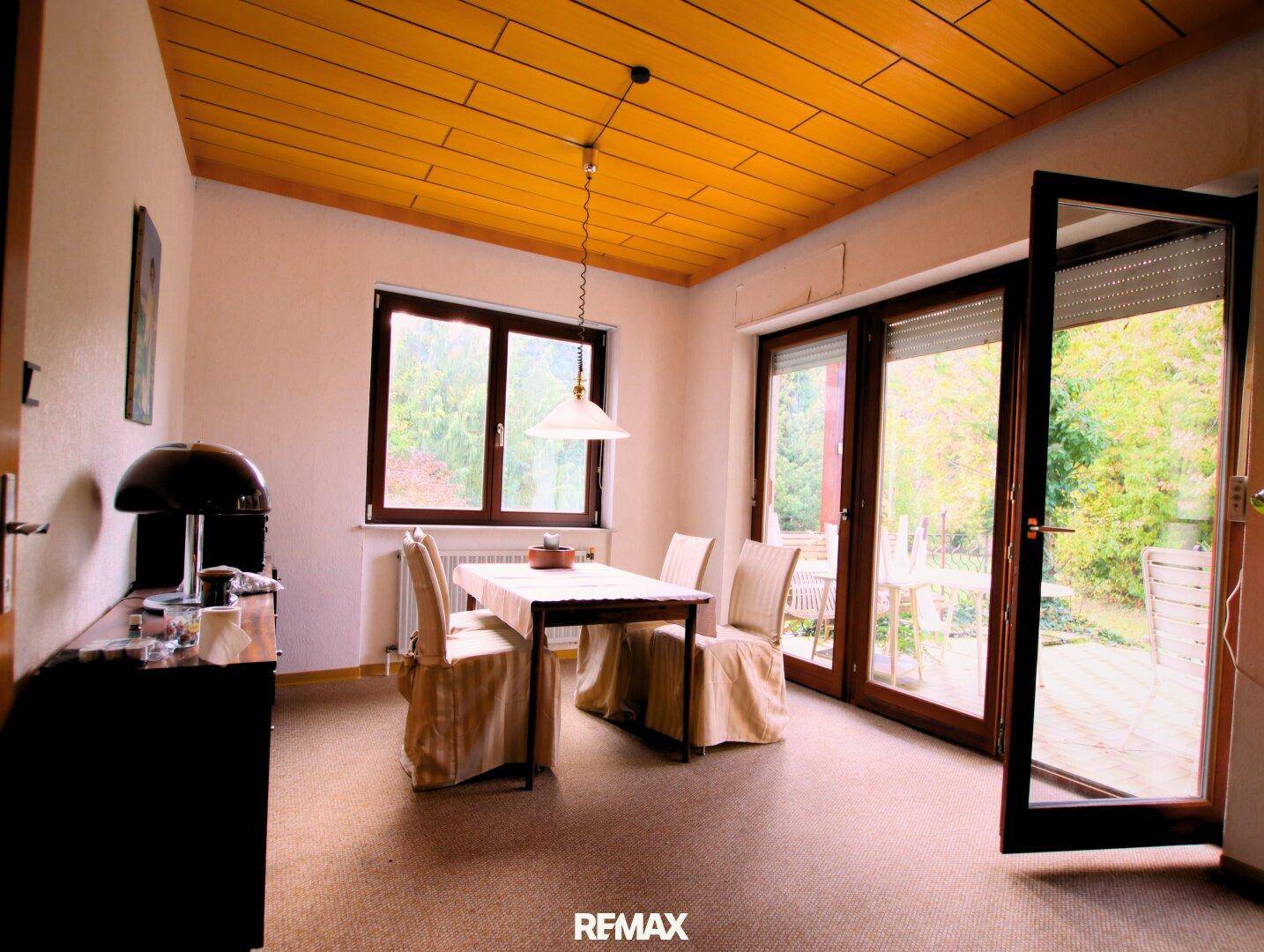 Esszimmer mit Holztisch, Stühlen und direktem Zugang zur Terrasse durch große Glastüren, unter einer Holzdecke.