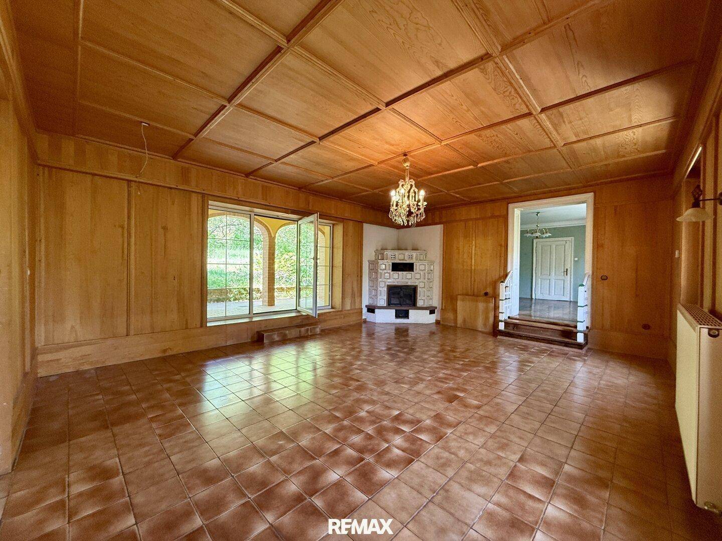 Geräumiges Wohnzimmer mit Holztäfelung, Kachelofen und Zugang zu einem Außenbereich.