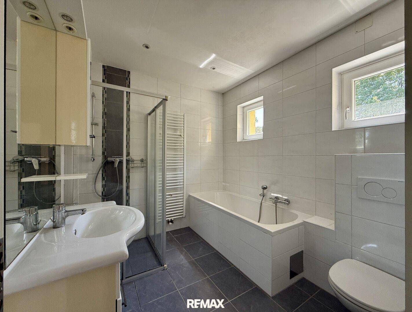 Modernes Badezimmer mit weißen Fliesen, Badewanne, Duschkabine und zwei Fenstern.