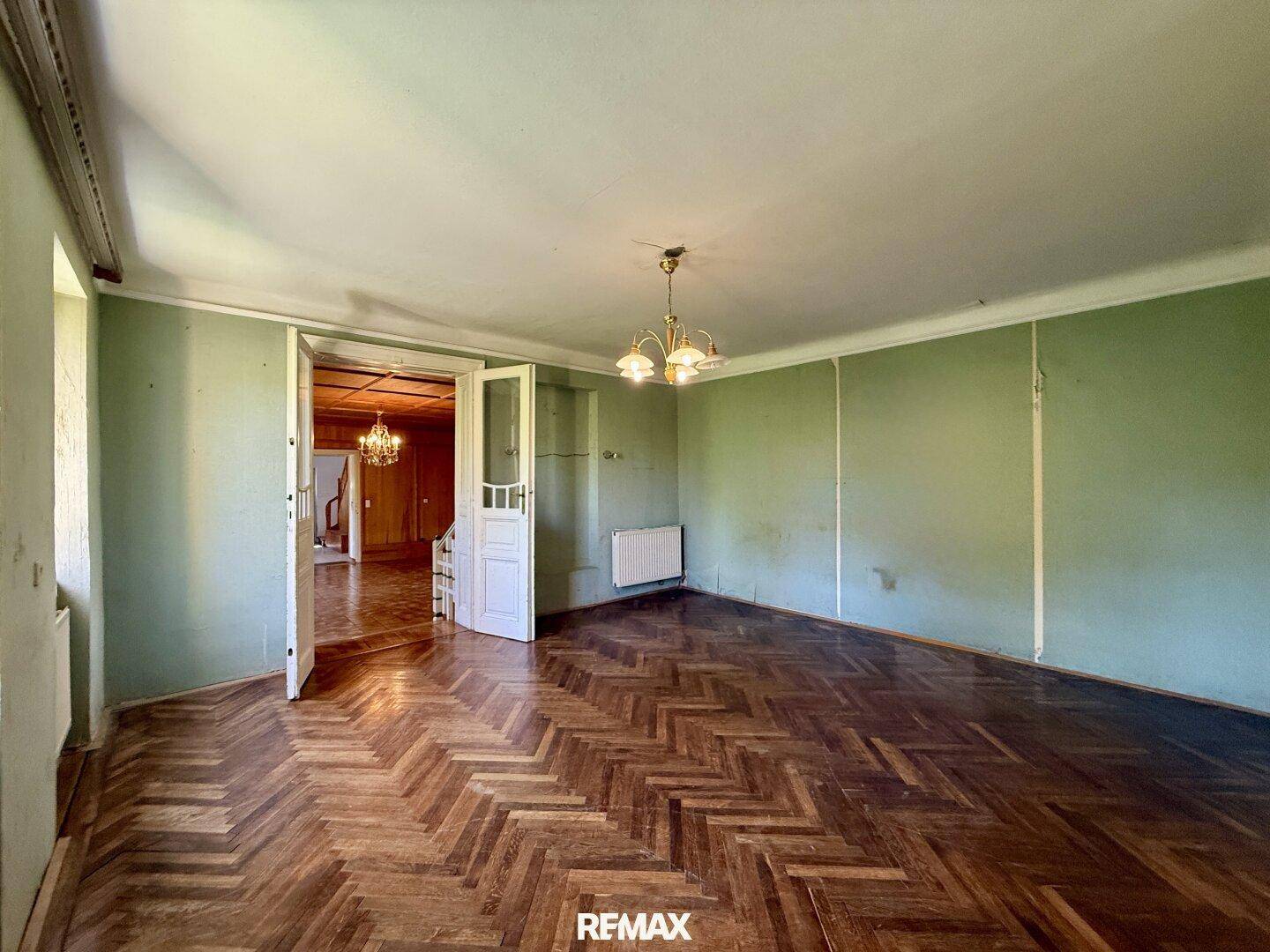 Geräumiges Zimmer mit grünen Wänden, Fischgrätparkett und Doppeltüren zu einem weiteren Raum.