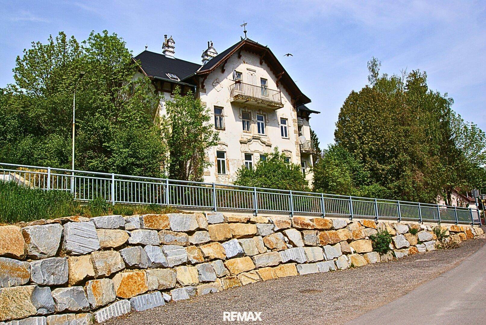Die Fassade des historischen Gebäudes mit Balkonen und einer Natursteinmauer im Vordergrund.