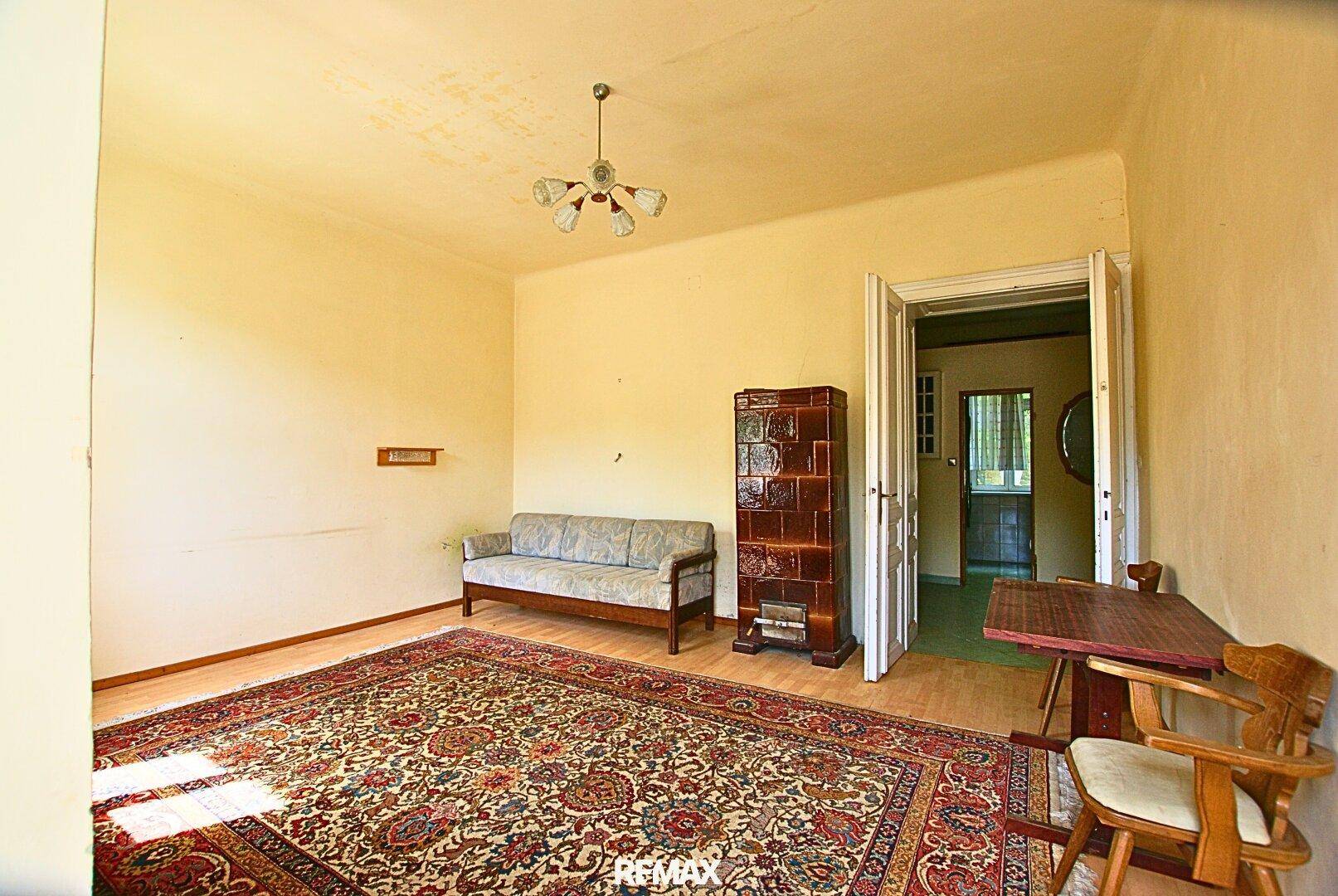 Gemütliches Wohnzimmer mit Kachelofen, Teppich und einfacher Einrichtung in einem Altbau.