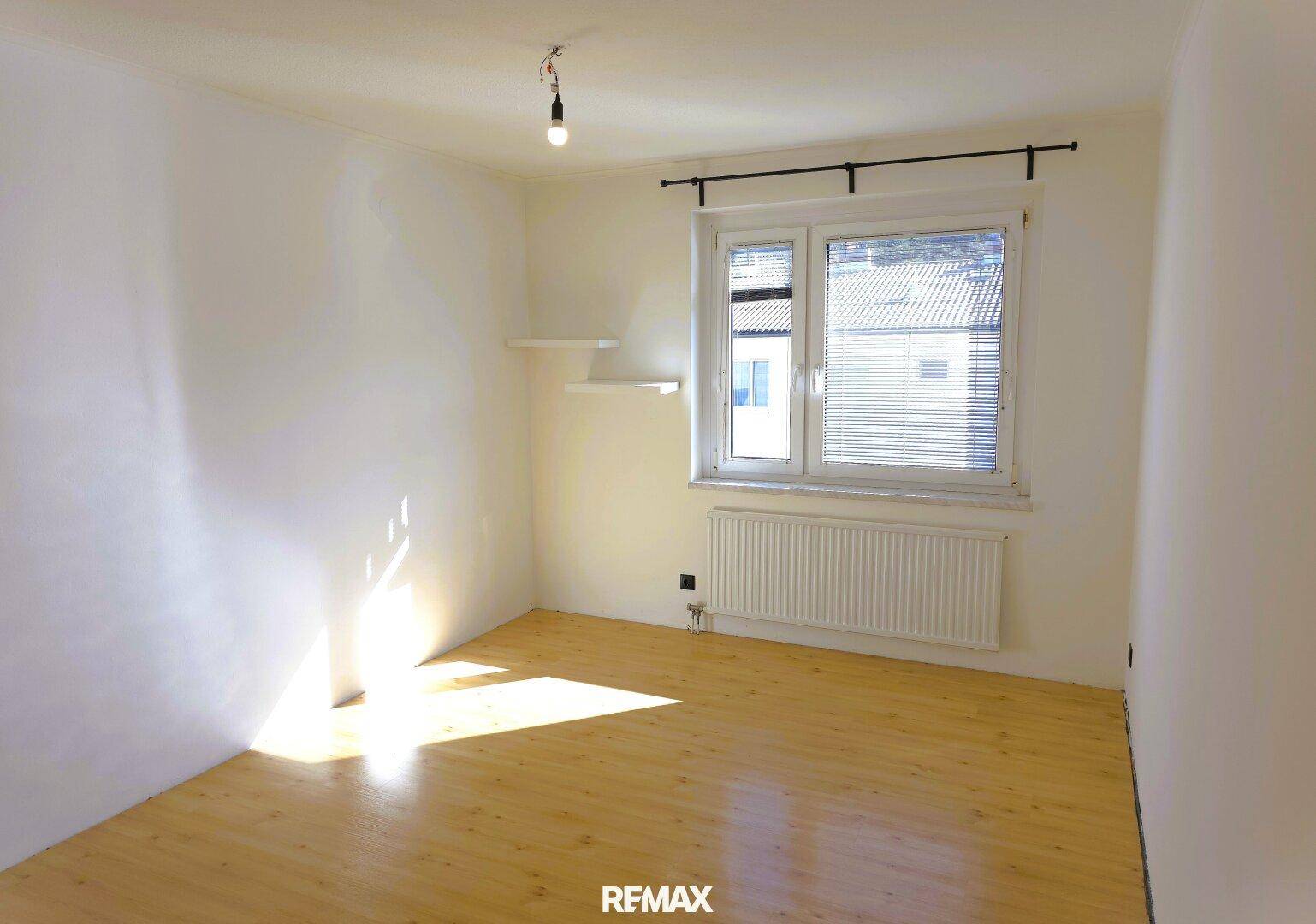 Helles, leeres Zimmer mit Holzfußboden, großem Fenster und Heizkörper.