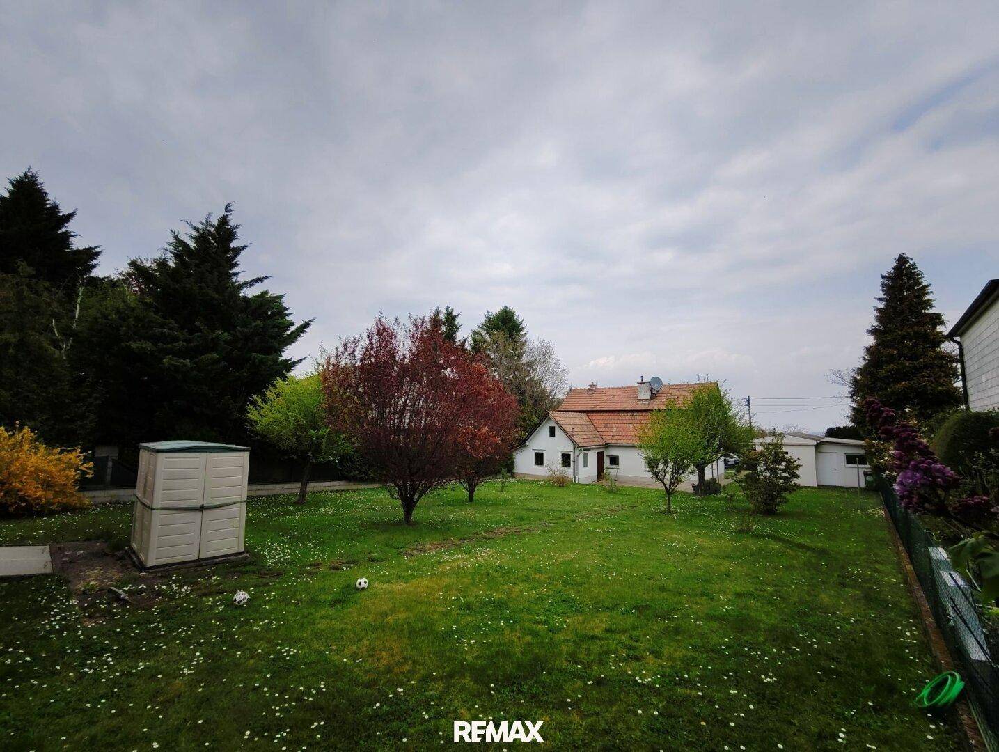 Großer, gepflegter Garten mit Rasenfläche, Bäumen und einem Gartenhaus im Hintergrund unter bewölktem Himmel.