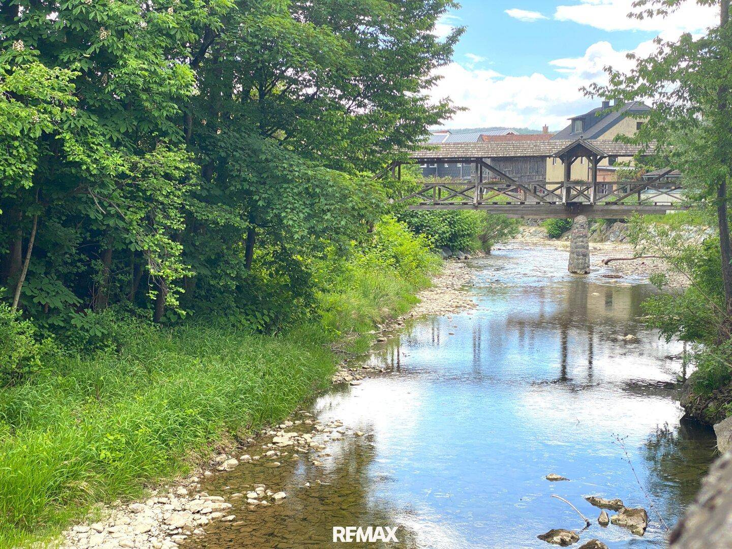 Idyllischer Flusslauf mit einer historischen Holzbrücke, umgeben von üppigem Grün.