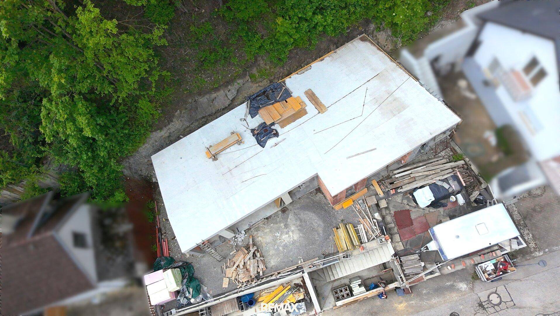 Luftaufnahme der Baustelle mit Flachdach und angrenzenden Gebäuden in grüner Umgebung.