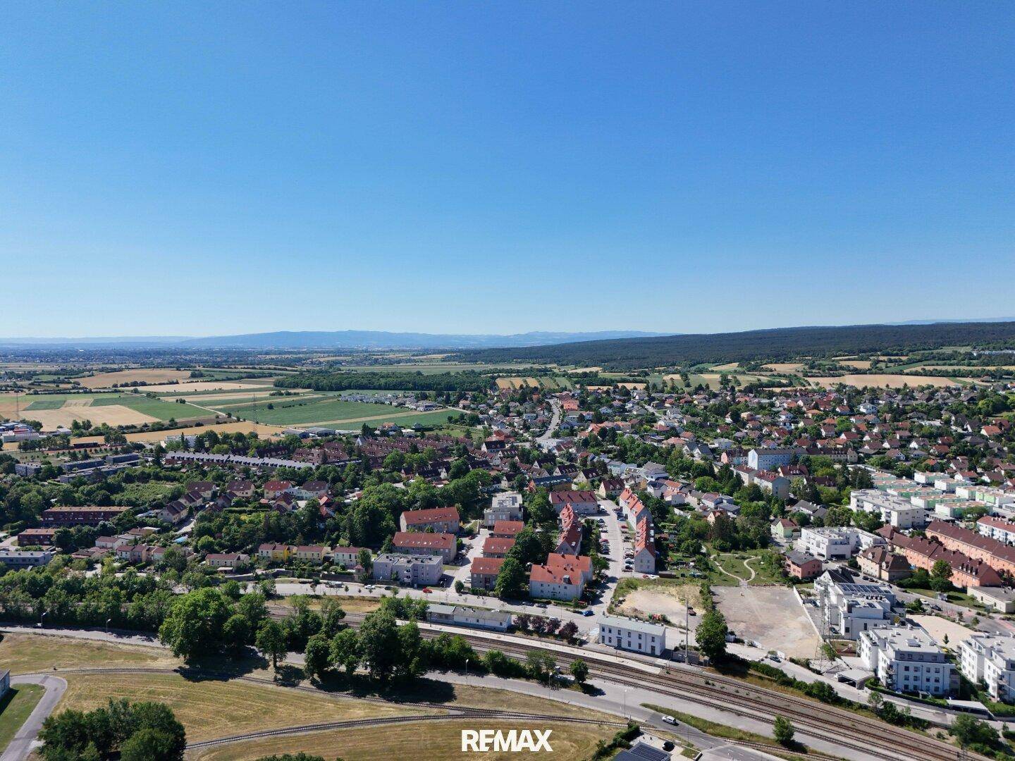Luftaufnahme einer Siedlung mit Wohnhäusern, Feldern und einer Bahnlinie, umgeben von grüner Landschaft.