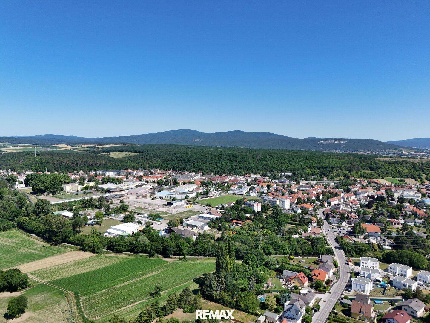 Weitläufige Luftaufnahme einer ländlichen Siedlung mit Häusern, Feldern und bewaldeten Hügeln im Hintergrund.