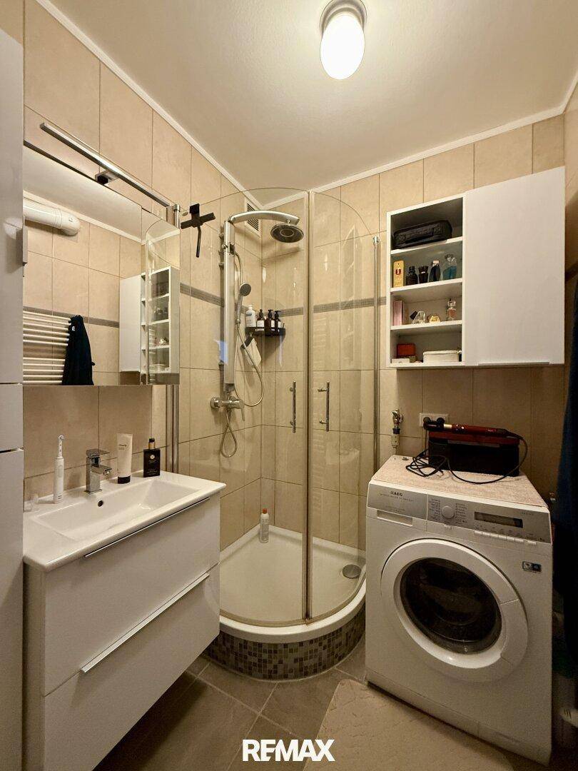 Gefliestes Badezimmer mit Duschkabine, Waschbecken und integrierter Waschmaschine.