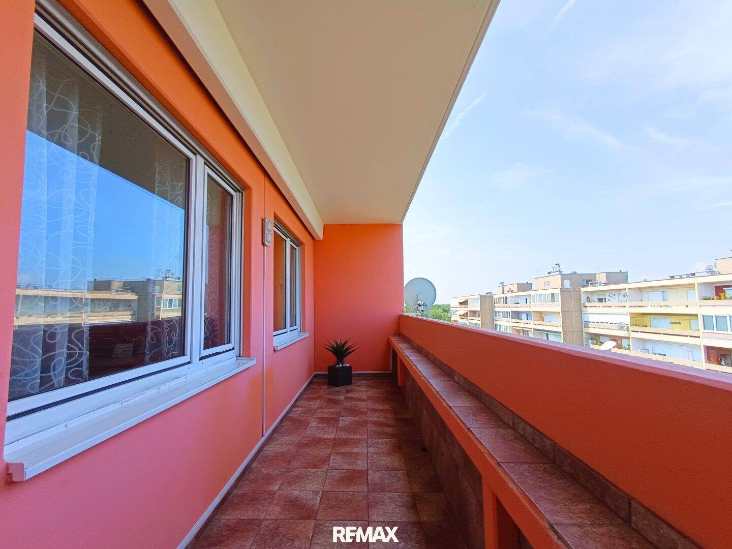 Langer Balkon mit orangefarbenen Wänden, Fliesenboden und Blick auf umliegende Gebäude.