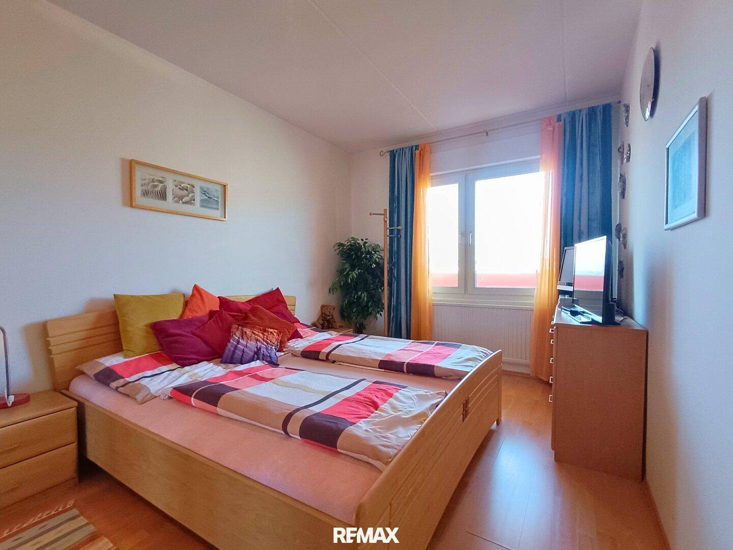 Schlafzimmer mit hellem Holzbett, farbenfroher Bettwäsche und großem Fenster.