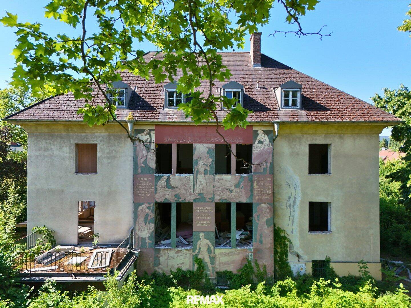 Fassade eines älteren Gebäudes mit Wandmalereien und sichtbaren Schäden, umgeben von Bäumen.