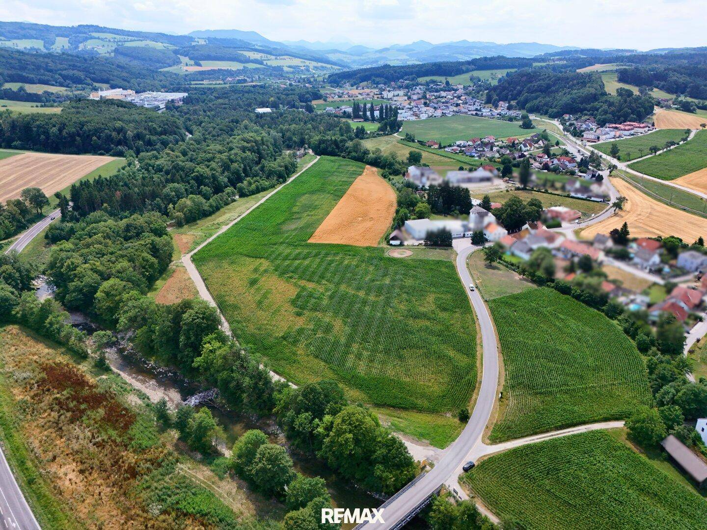 Weitläufige Luftaufnahme einer hügeligen Landschaft mit Feldern, Wäldern, einem Fluss und einem Dorf in der Ferne.