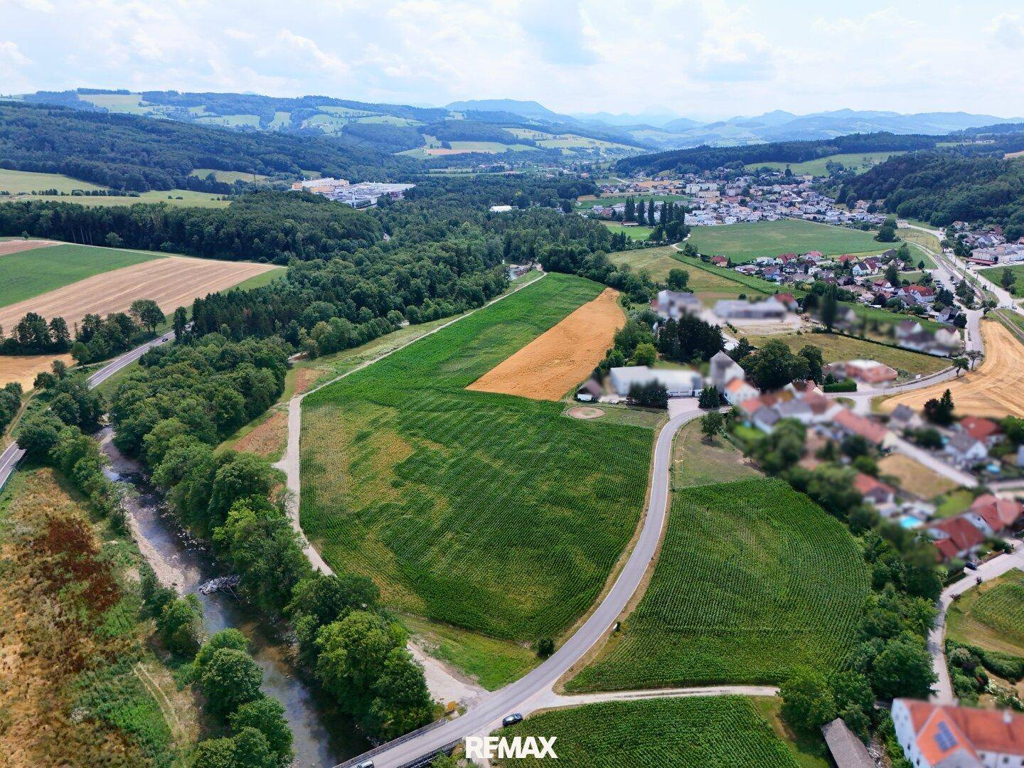 Weitläufige Luftaufnahme einer hügeligen Landschaft mit Feldern, Wäldern, einem Fluss und einem Dorf in der Ferne.