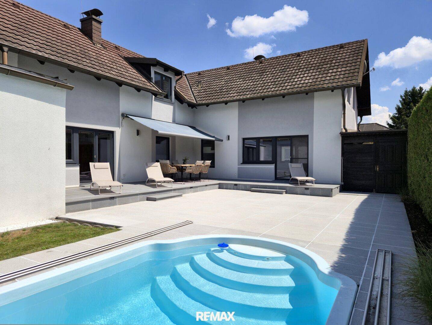 Blick auf den Poolbereich und die Terrasse mit Sonnenliegen hinter dem modernen Haus.