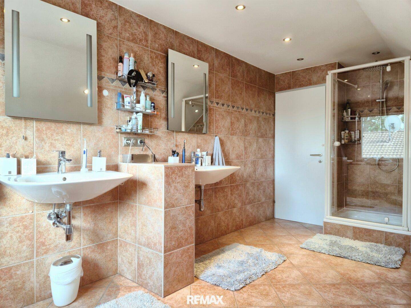 Badezimmer mit zwei Waschbecken, Duschkabine und braunen Fliesen.