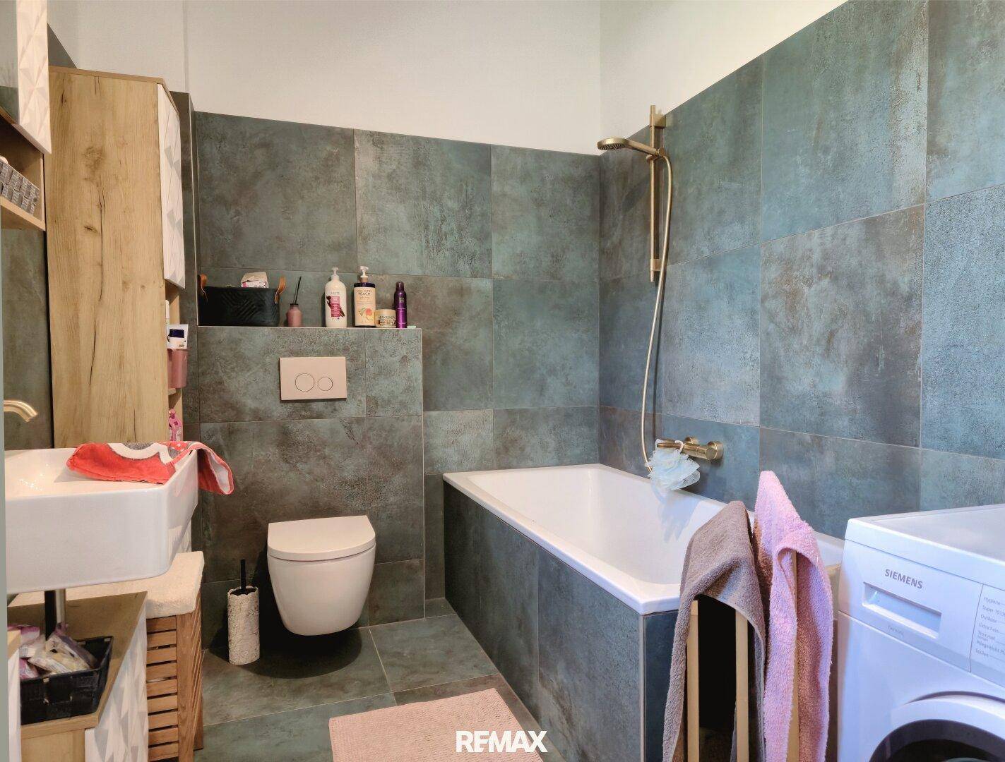 Badezimmer mit dunklen Fliesen, Badewanne, Wand-WC und Waschbecken.