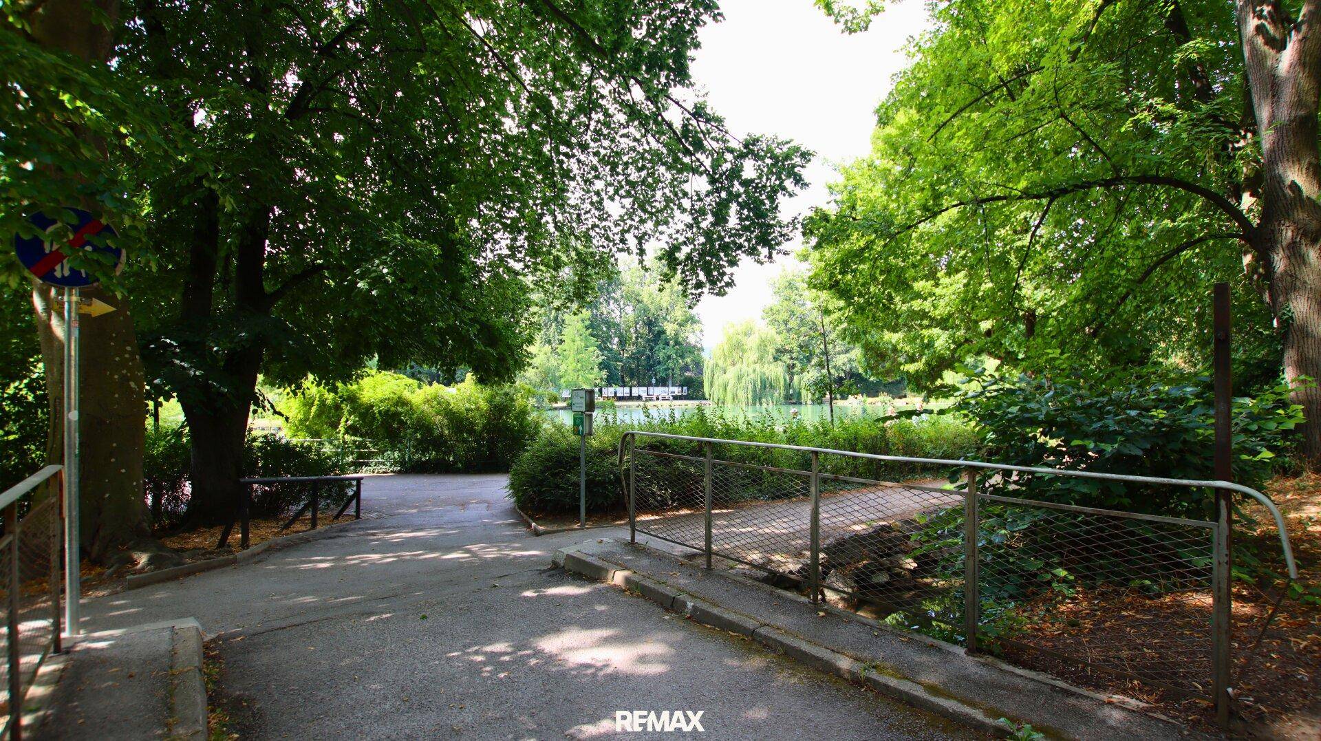 Spazierweg führt über eine kleine Brücke zu einem idyllischen See, umgeben von Bäumen.
