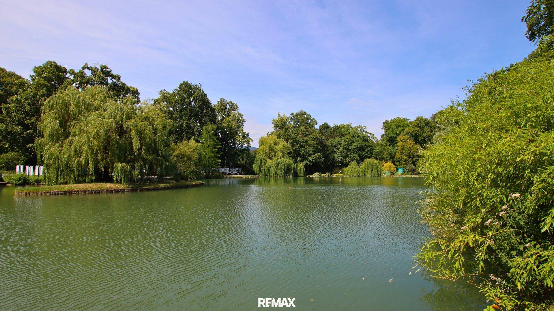 Weitläufiger See mit kleiner Insel, umgeben von dichtem Grün unter blauem Himmel.
