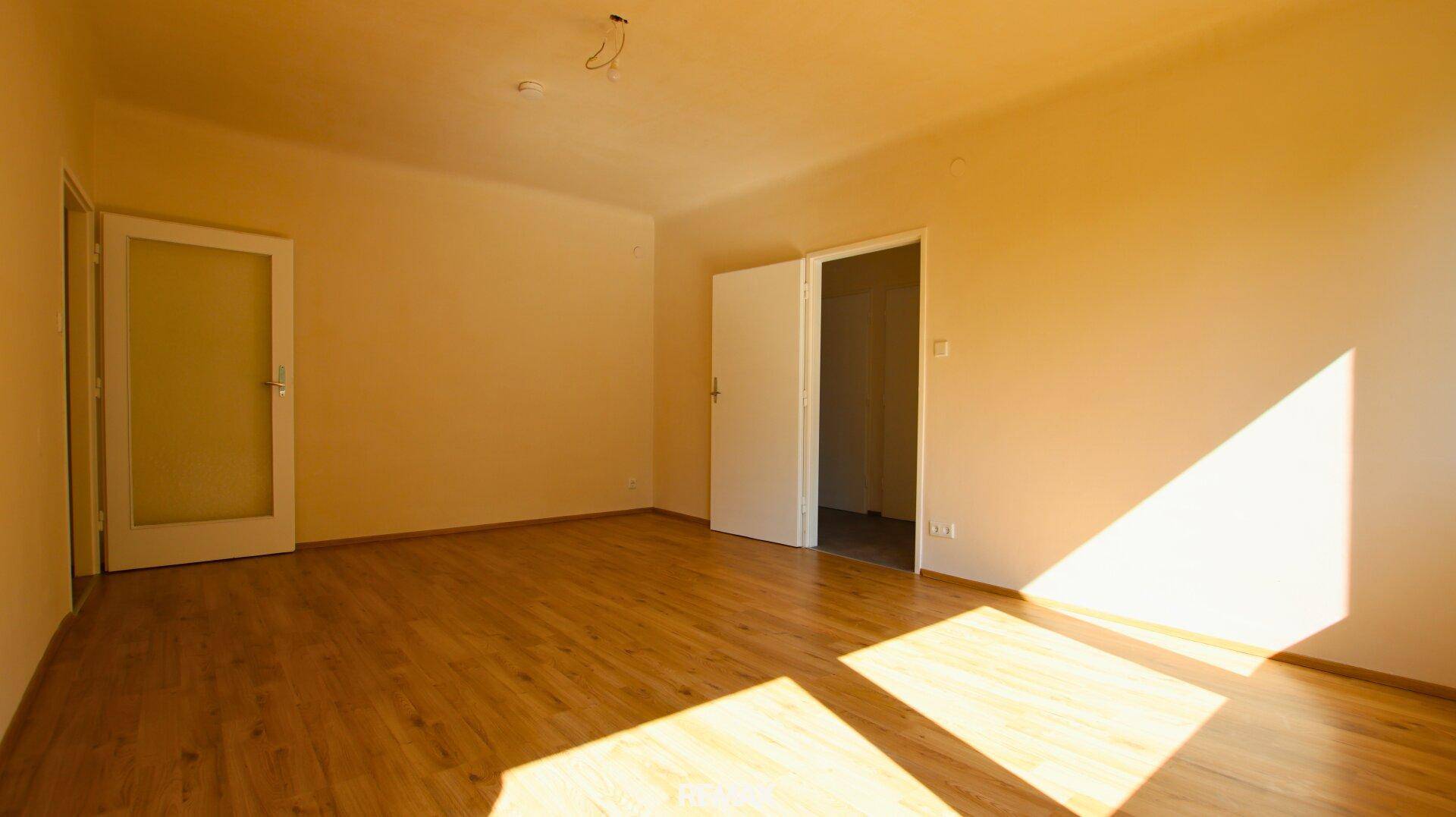 Leerer Raum mit Holzboden und hellen Wänden, durchflutet von Sonnenlicht.