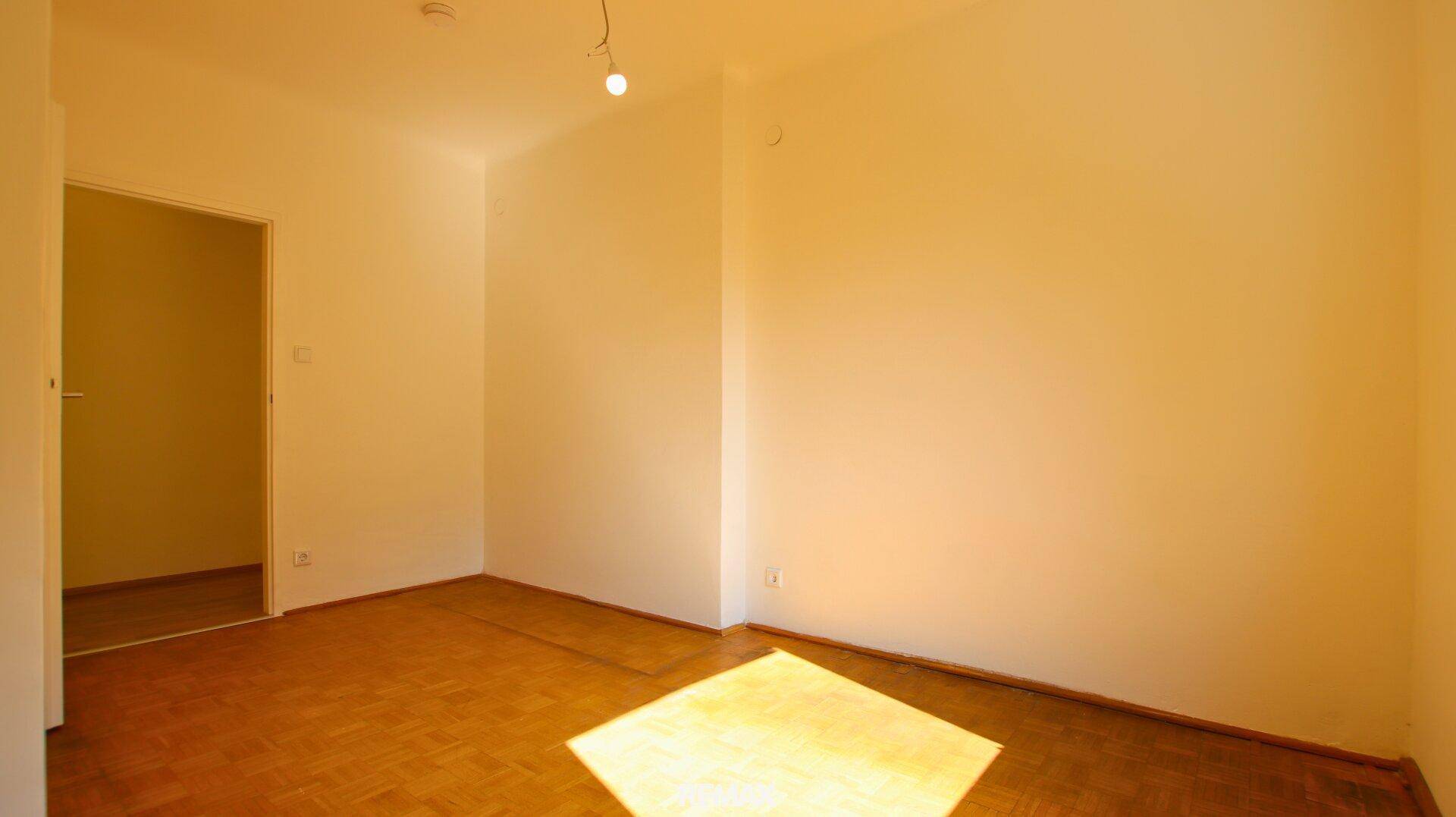 Leeres Schlafzimmer mit Parkettboden und hellen Wänden, durchflutet von Sonnenlicht.