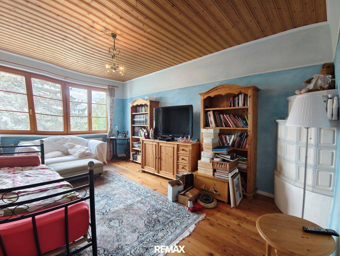 Helles Zimmer mit Holzbalkendecke, Kachelofen und blauen Wänden, als Schlaf- oder Wohnraum nutzbar.