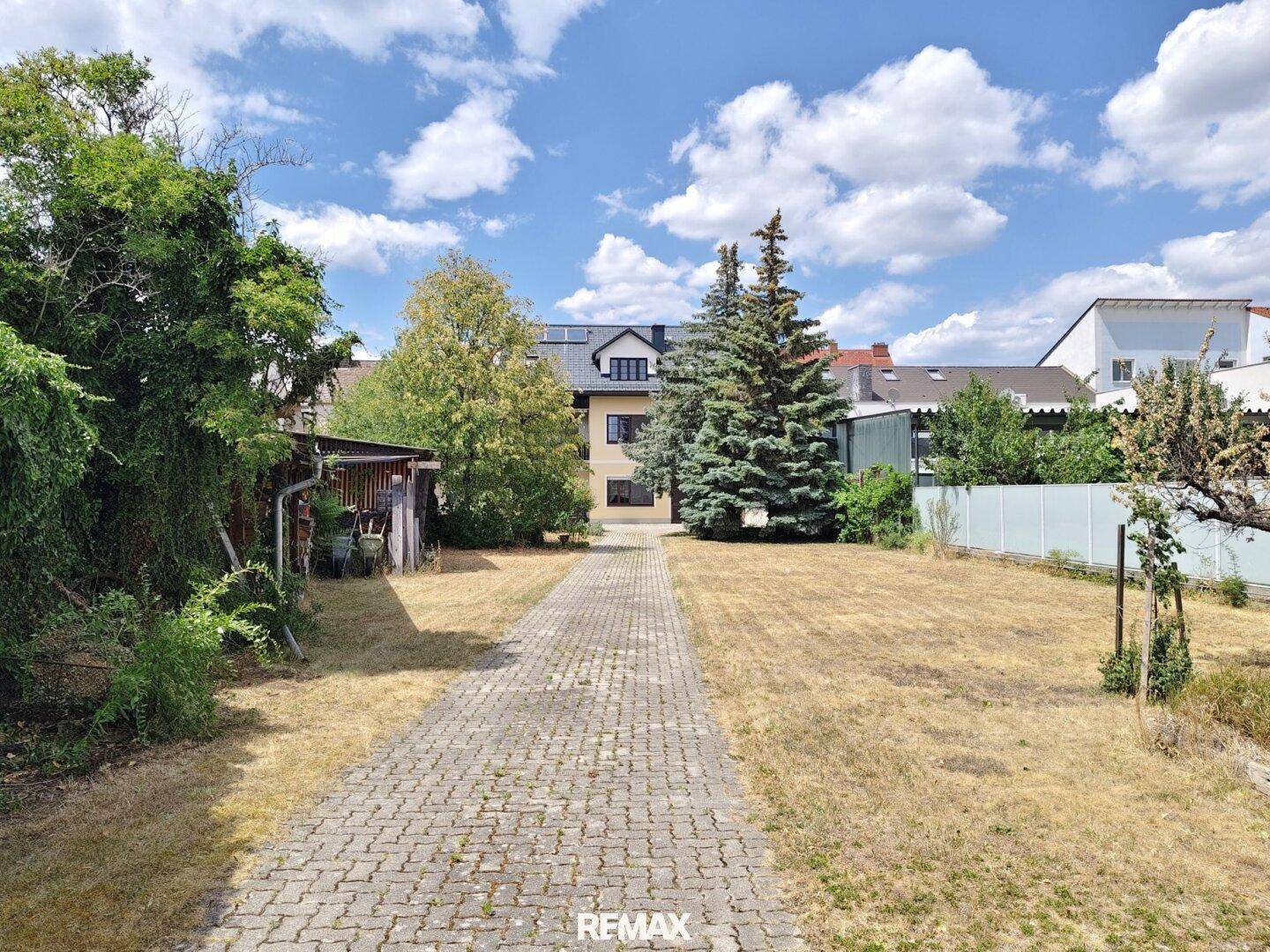 Großer Gartenbereich mit gepflastertem Weg, Rasenfläche und altem Baumbestand im Hintergrund.