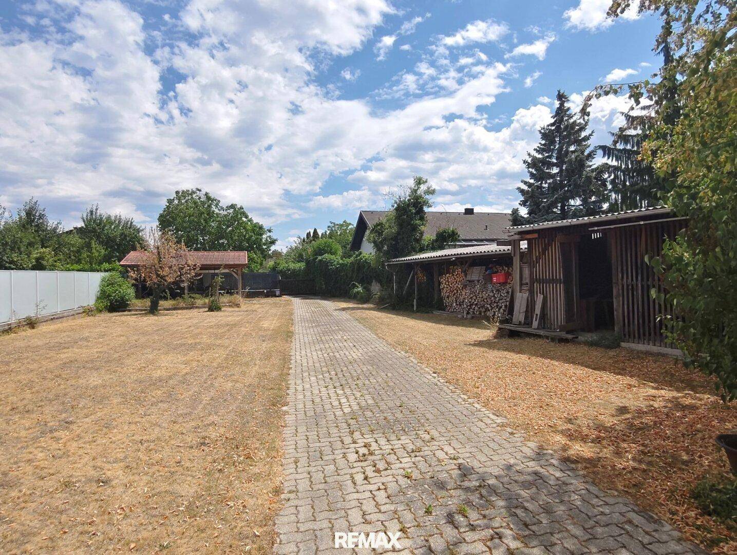 Langer gepflasterter Weg durch den Garten mit Holzschuppen und Brennholzstapel.