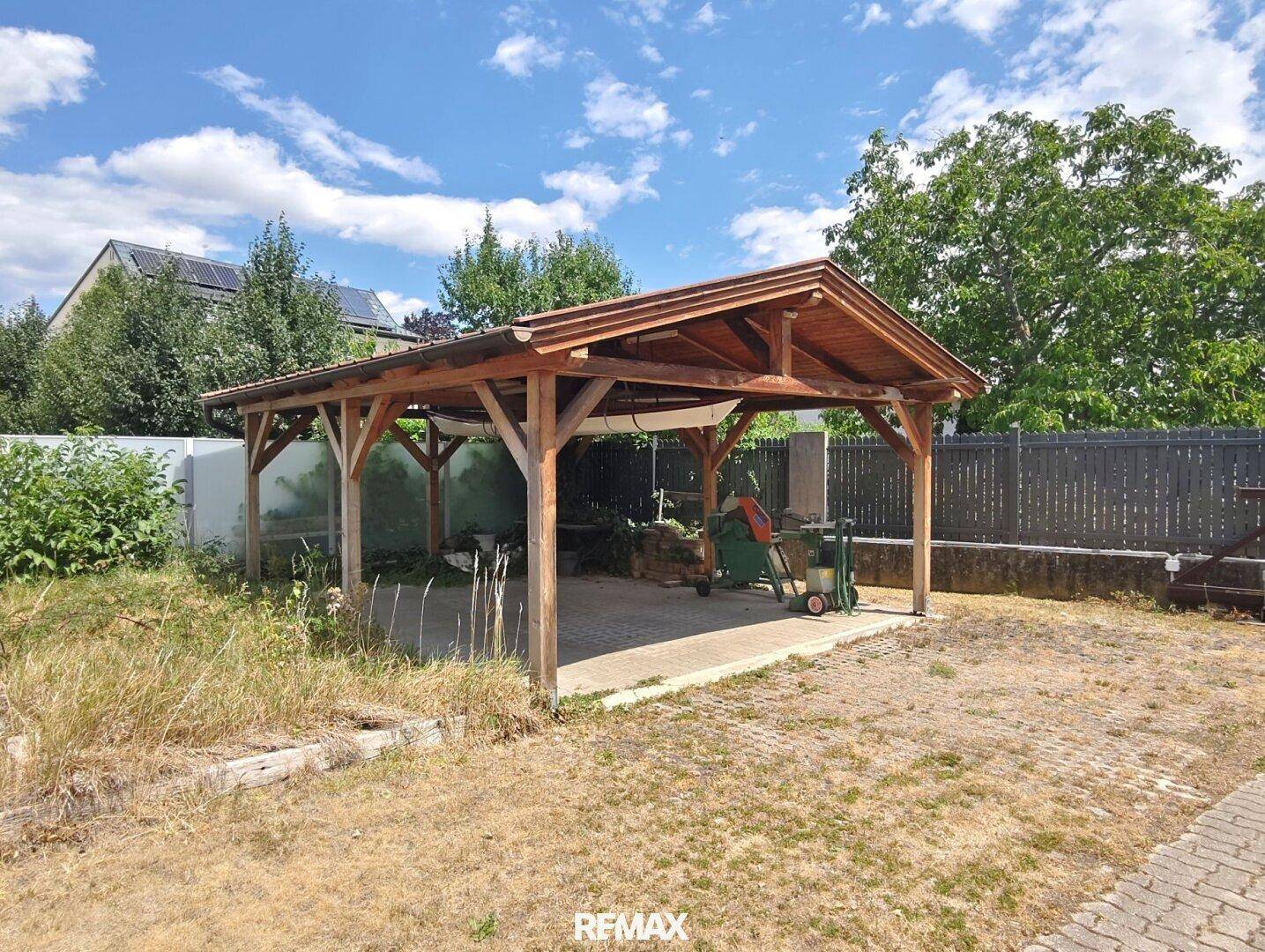 Offener Holzcarport auf gepflastertem Bereich im Garten.