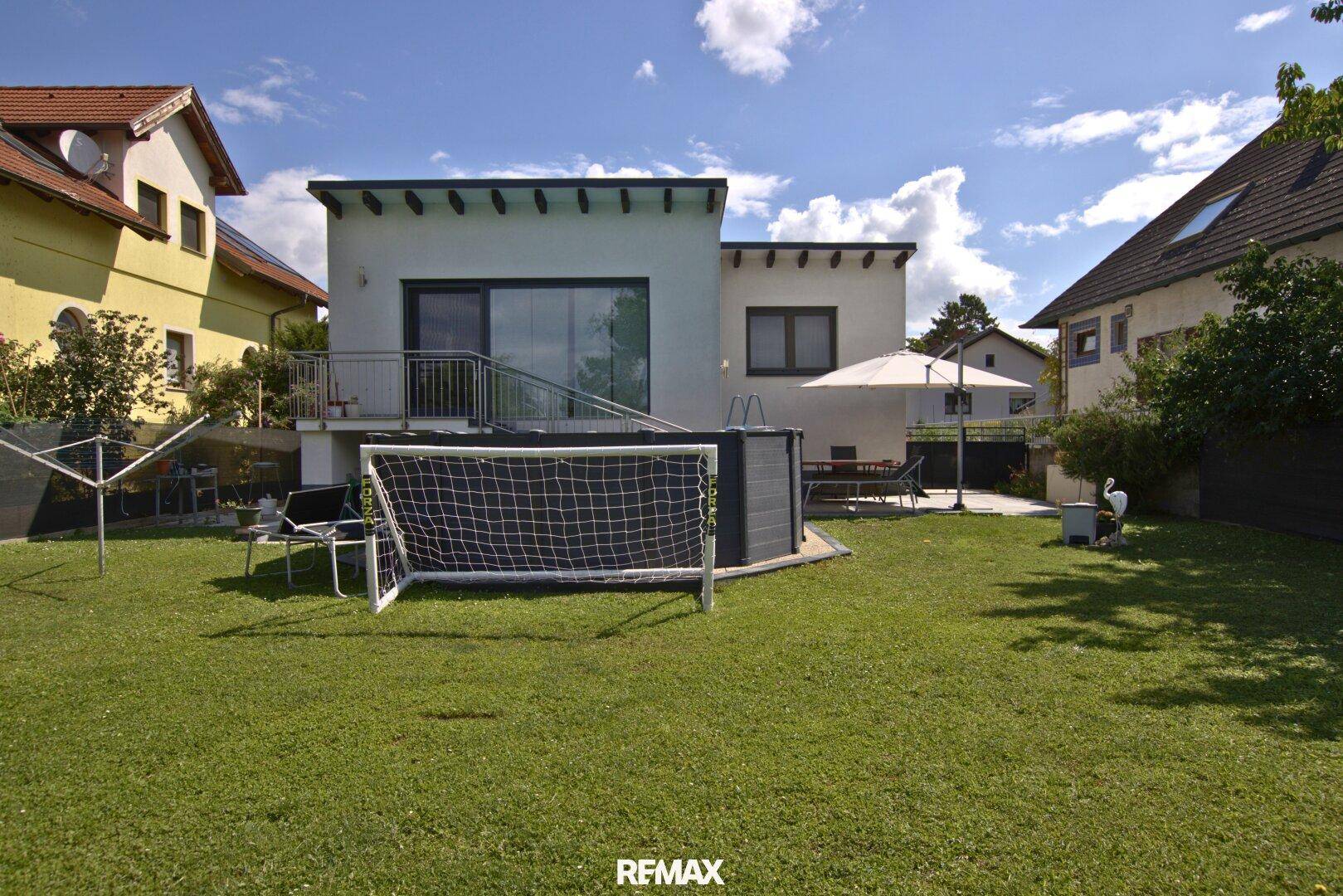Rückansicht des Hauses mit Garten, Pool und Terrasse unter blauem Himmel.