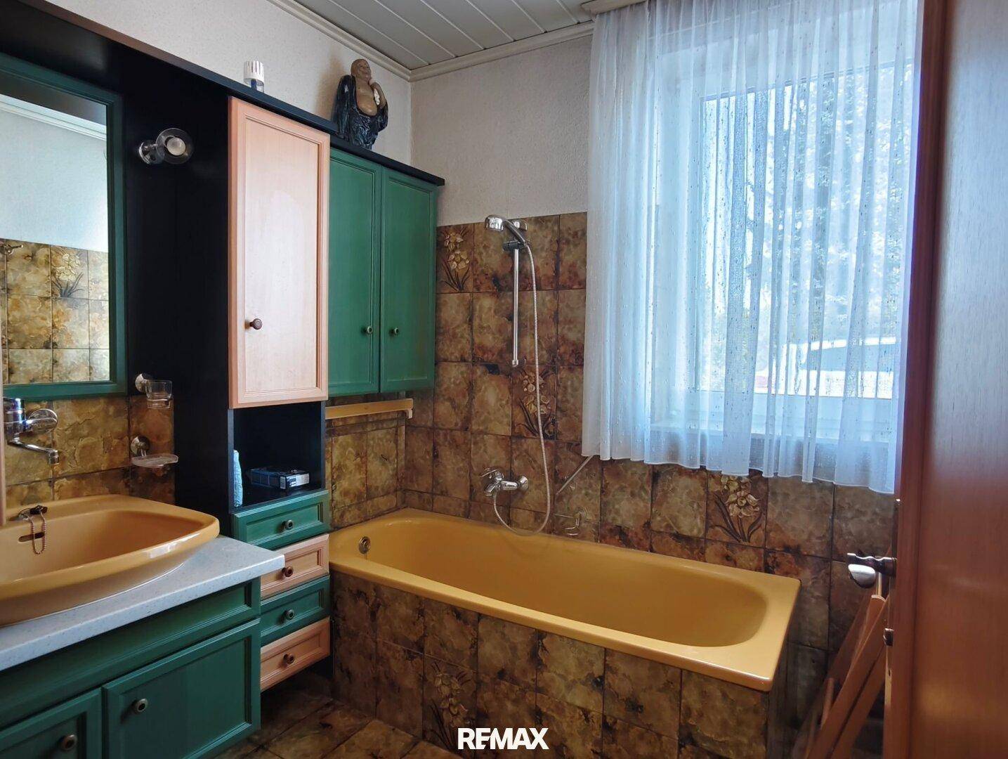 Badezimmer im Stil der 80er Jahre mit gelber Badewanne, Dusche, Waschbecken und gemusterten Fliesen.