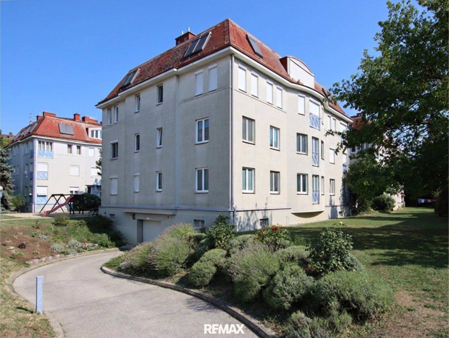 Mehrfamilienhaus mit rotem Mansarddach, umgeben von einer gepflegten Grünanlage.