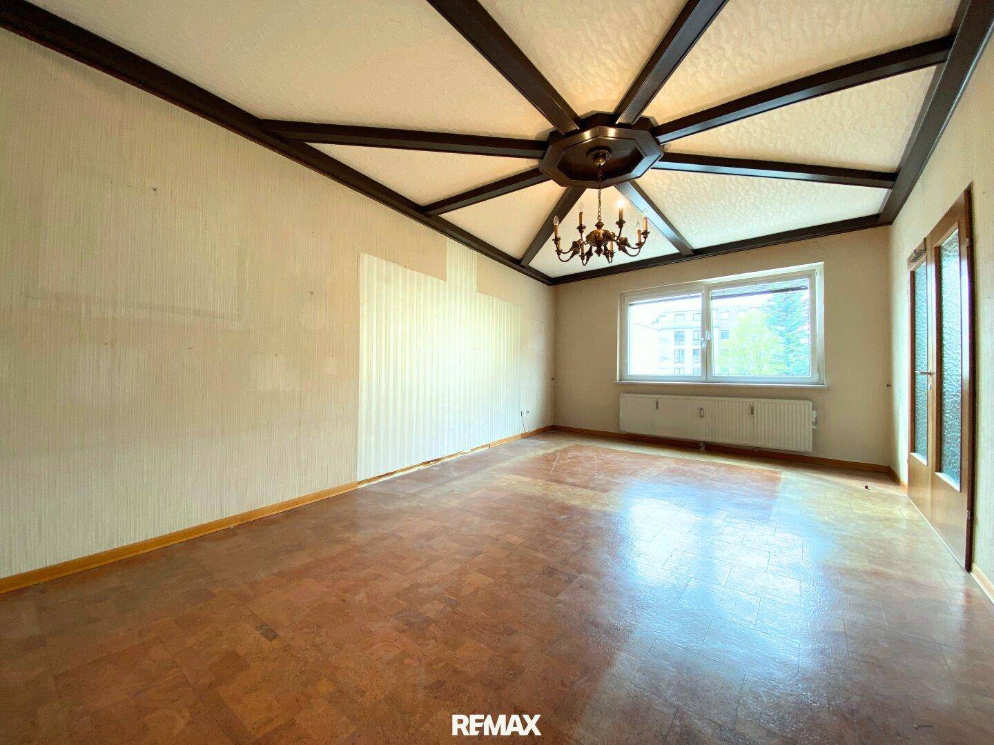 Geräumiges Wohnzimmer mit Parkettboden, dekorativer Holzdecke und Kronleuchter, bereit zur Renovierung.