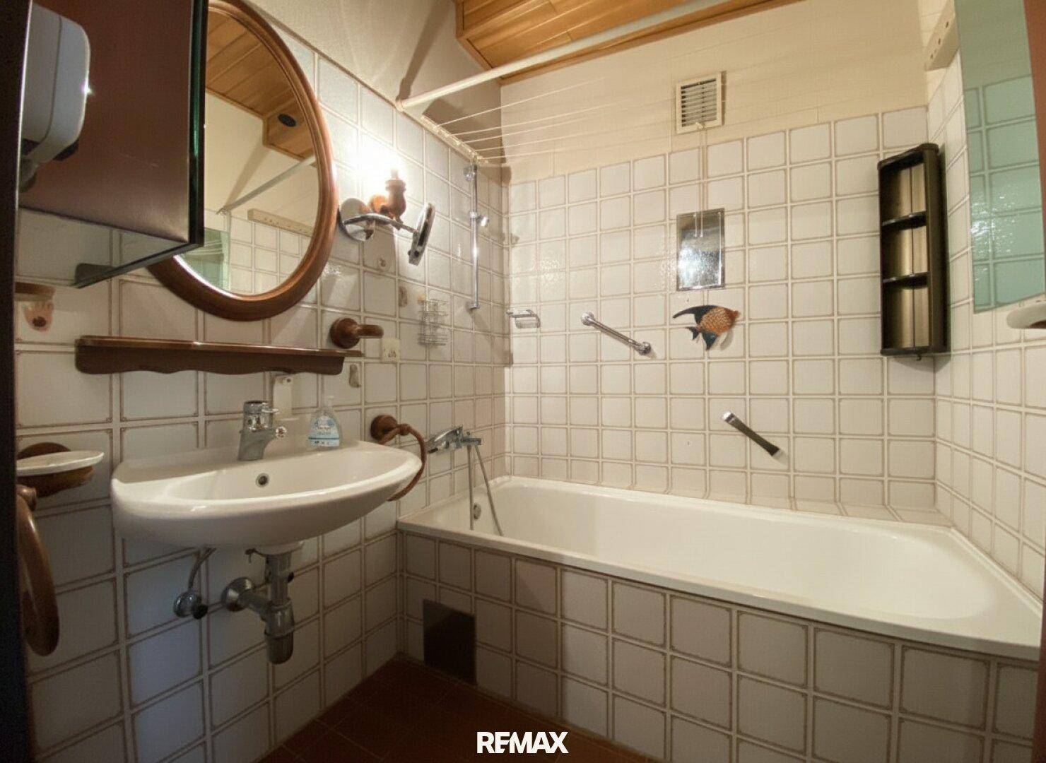 Badezimmer mit weißen Fliesen, Badewanne mit Duschfunktion und einem Waschbecken unter Holzdecke.