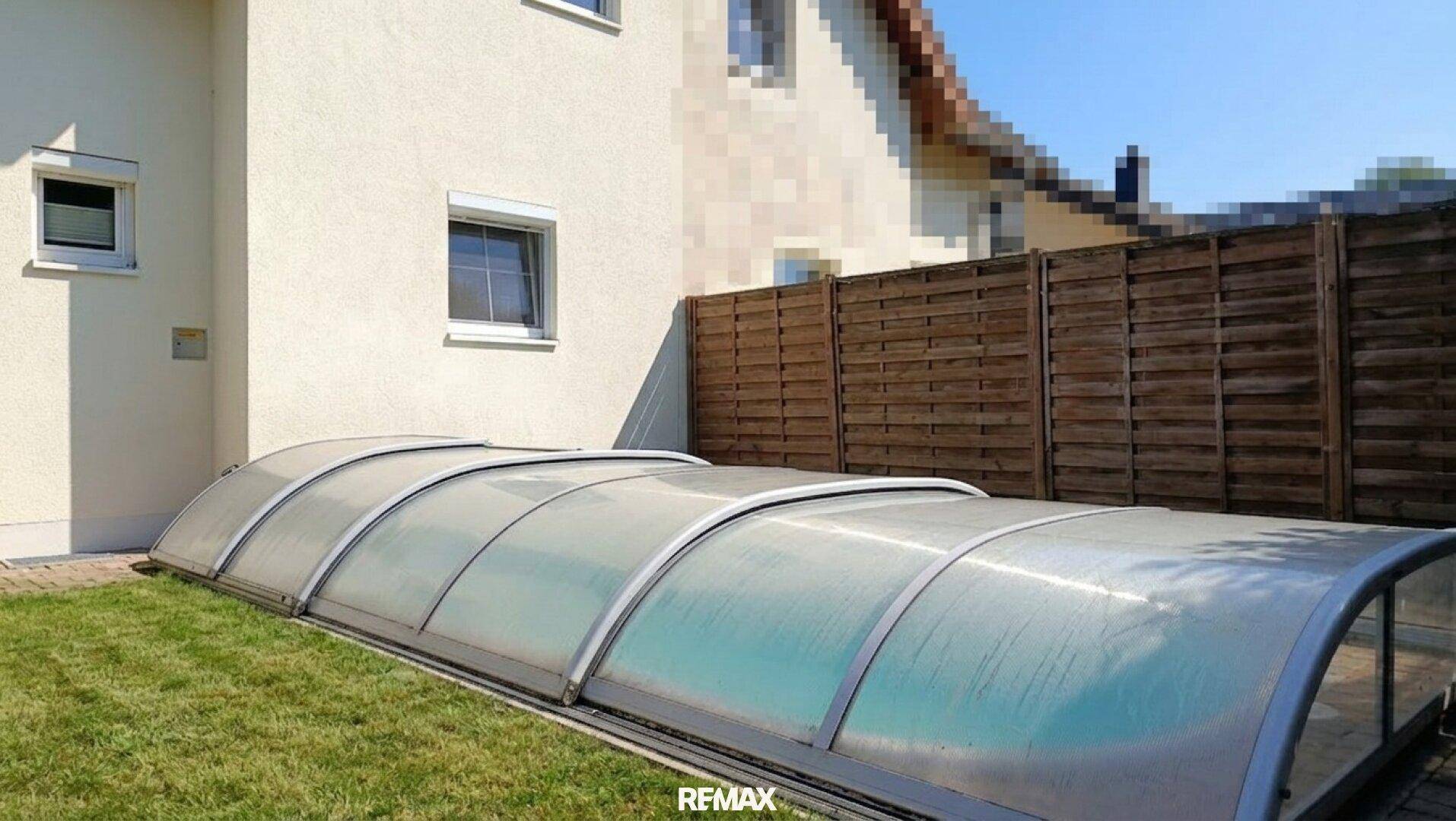 Der abgedeckte Swimmingpool im Garten, angrenzend an die Hausfassade und einen Holzzaun.