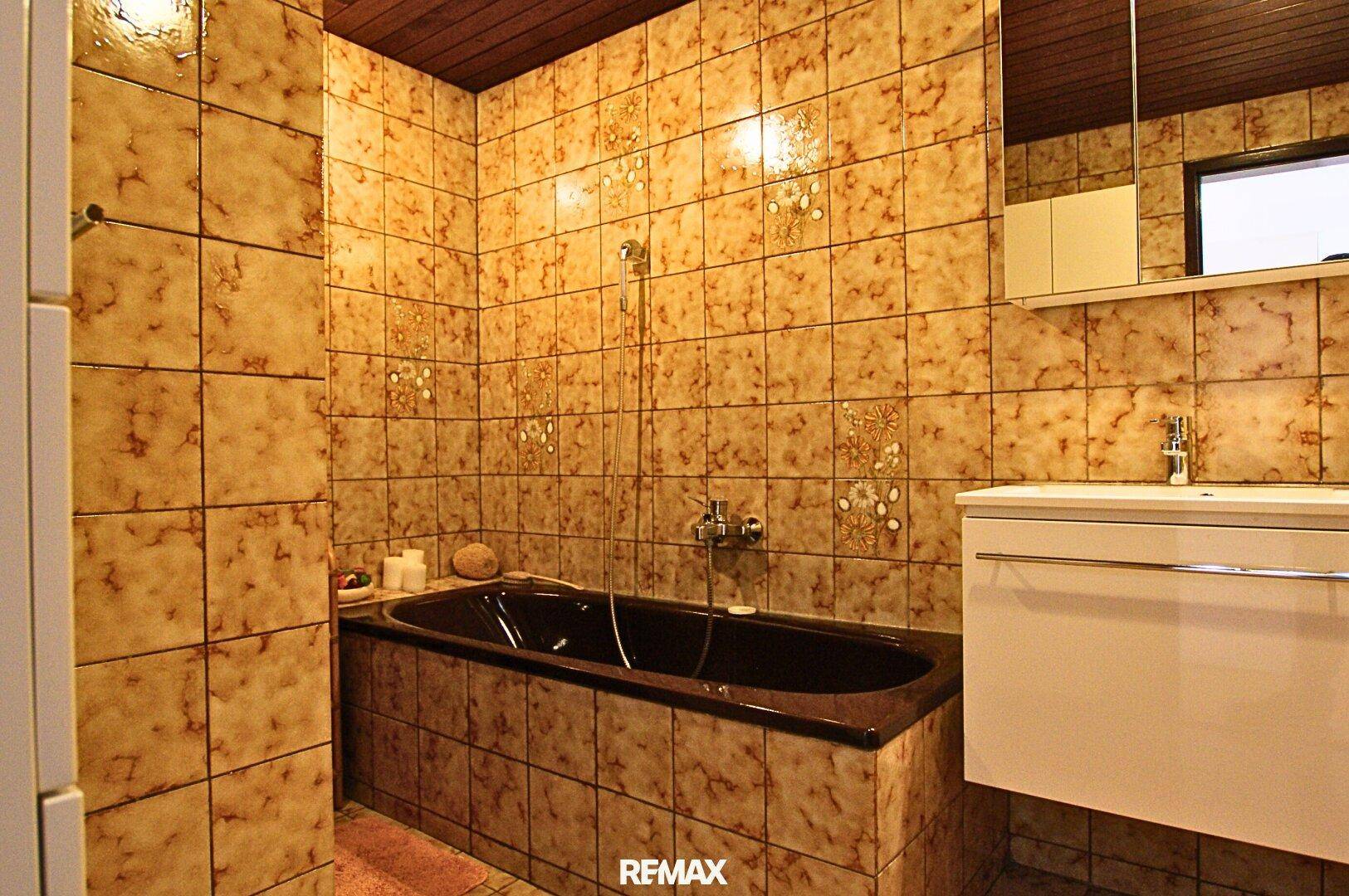 Badezimmer mit gemusterten Fliesen, dunkler Badewanne, modernem Waschtisch und Holzdecke.