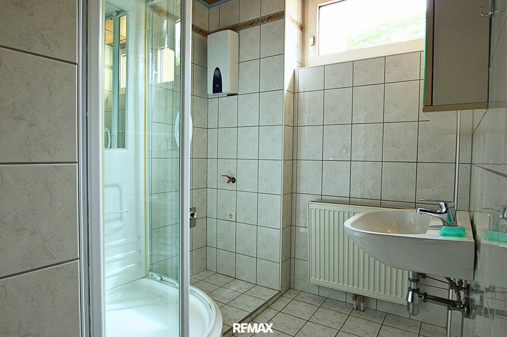 Badezimmer mit weiß gefliesten Wänden, Eckdusche, Waschbecken und einem Fenster für Tageslicht.