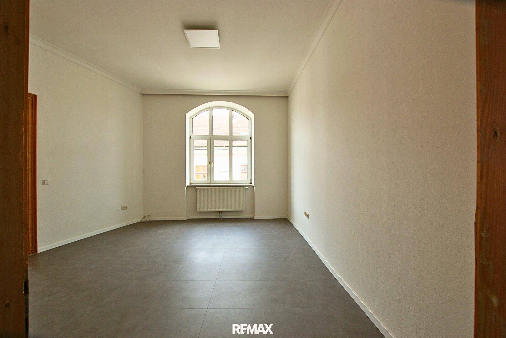Leerer Raum mit grauen Fliesen, weißen Wänden und einem großen Bogenfenster.