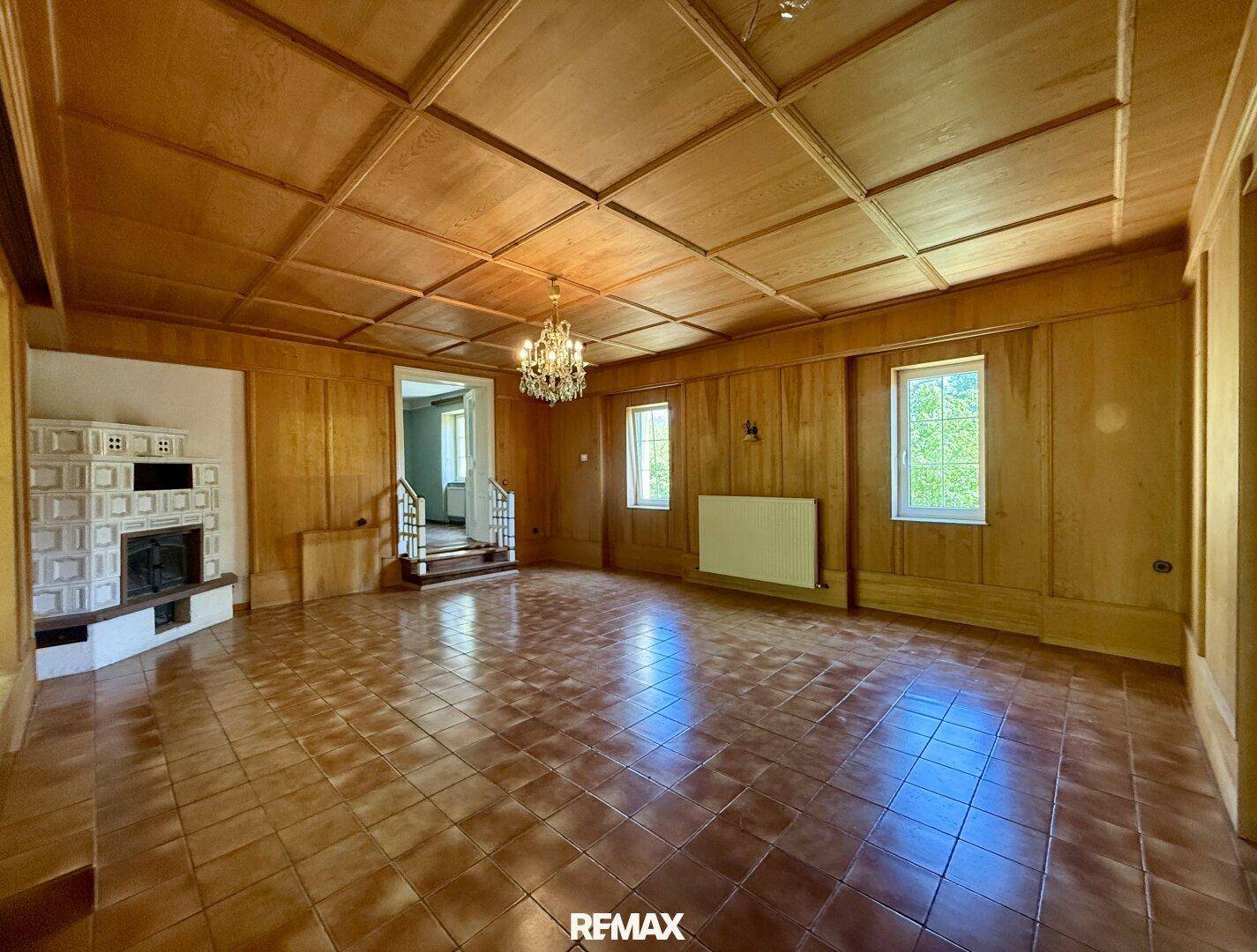 Geräumiges Wohnzimmer mit Holztäfelung, Kachelofen und Terrakottafliesen, bereit zur Renovierung.