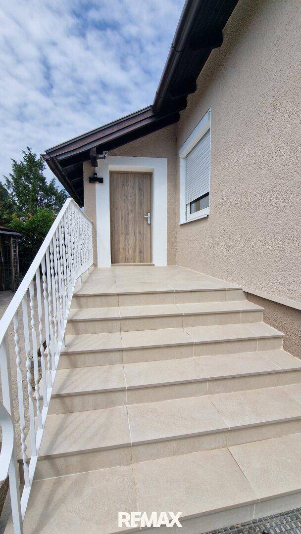 Eingangsbereich des Hauses mit gefliester Treppe, weißem Geländer und Holztür.