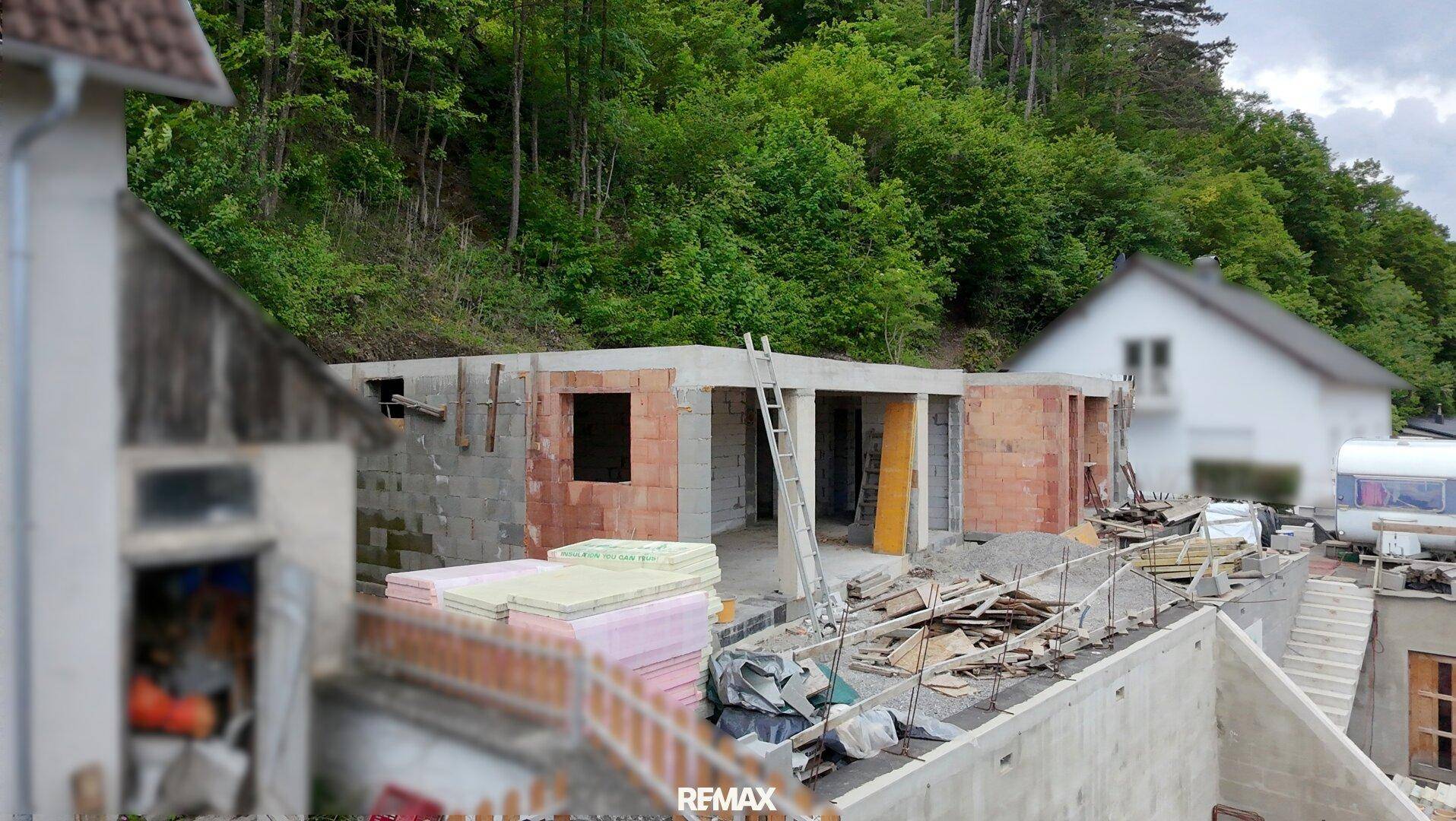 Baustelle eines neuen Hauses mit Rohbauwänden und Baumaterialien, umgeben von Bäumen.