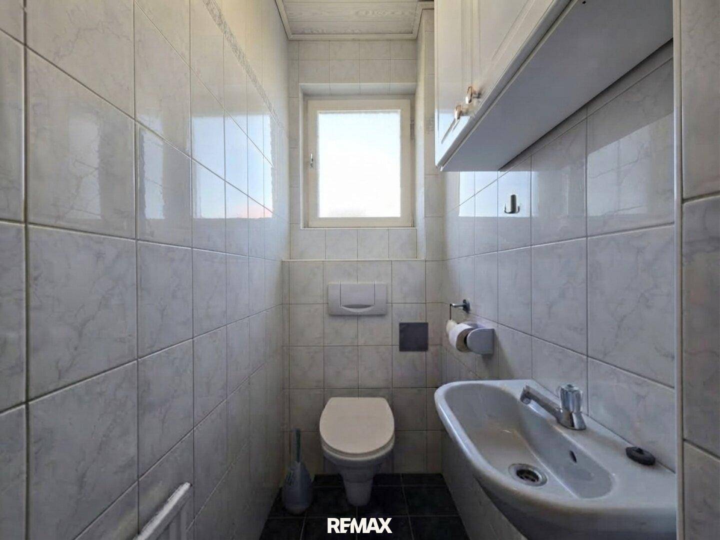 Separates WC mit Toilette, Waschbecken und hellen Wandfliesen, ausgestattet mit Fenster.