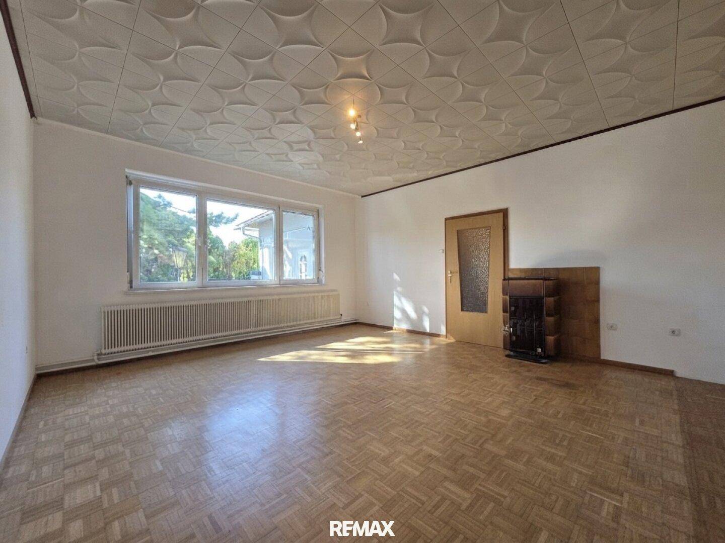 Helles Wohnzimmer mit Parkettboden, großem Fenster und traditionellem Kachelofen.