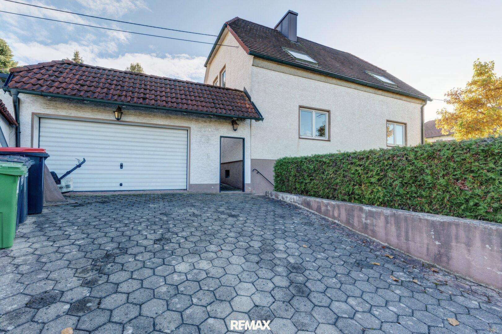Außenansicht eines Hauses mit integrierter Garage, gepflasterter Einfahrt und Hecke.